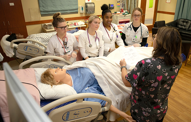 Health Sciences students around mannequin listening to instructor